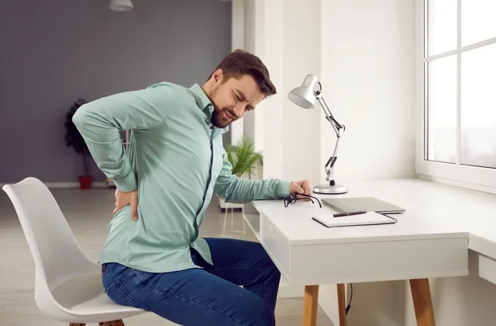 Zdjęcie przedstawia mężczyzne cierpiącego z bólu, który siedzi przy biurku na niewygodnym krześle