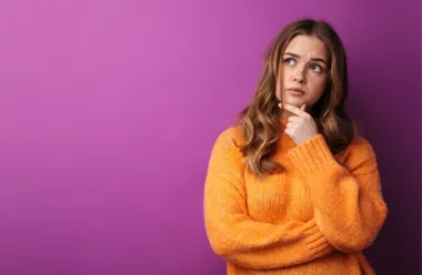 Młoda kobieta w pomarańczowym swetrze, zamyślona, na fioletowym tle.