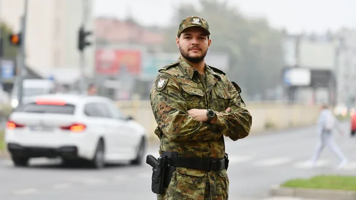Funkcjonariusz w polskim mundurze wojskowym stoi z założonymi rękami na ulicznym skrzyżowaniu, w tle przejeżdża biały samochód.