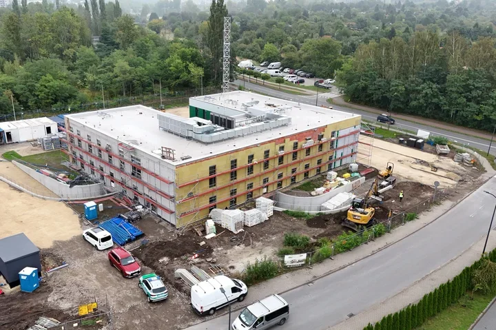 Widok z lotu ptaka na budowę wielokondygnacyjnego budynku przy ulicy, z koparkami, materiałami budowlanymi i zaparkowanymi pojazdami na placu.