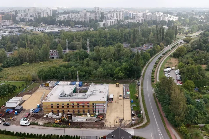 Widok z lotu ptaka na budowę nowego obiektu przy krętej drodze, z zabudową wielorodzinną i lasem w tle.