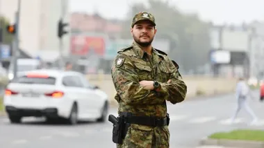 Funkcjonariusz w polskim mundurze wojskowym stoi z założonymi rękami na ulicznym skrzyżowaniu, w tle przejeżdża biały samochód.