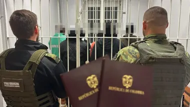 Funkcjonariusze Straży Granicznej pilnują osób zatrzymanych za kratami, na pierwszym planie widoczne są dwa paszporty Kolumbii.
