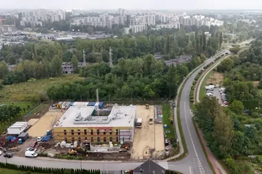 Widok z lotu ptaka na budowę nowego obiektu przy krętej drodze, z zabudową wielorodzinną i lasem w tle.