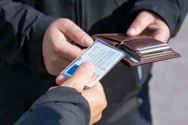 Dwie osoby w ciemnych kurtkach wymieniają dokument tożsamości w przezroczystej okładce, jedna z nich trzyma otwarty brązowy portfel z kartami.